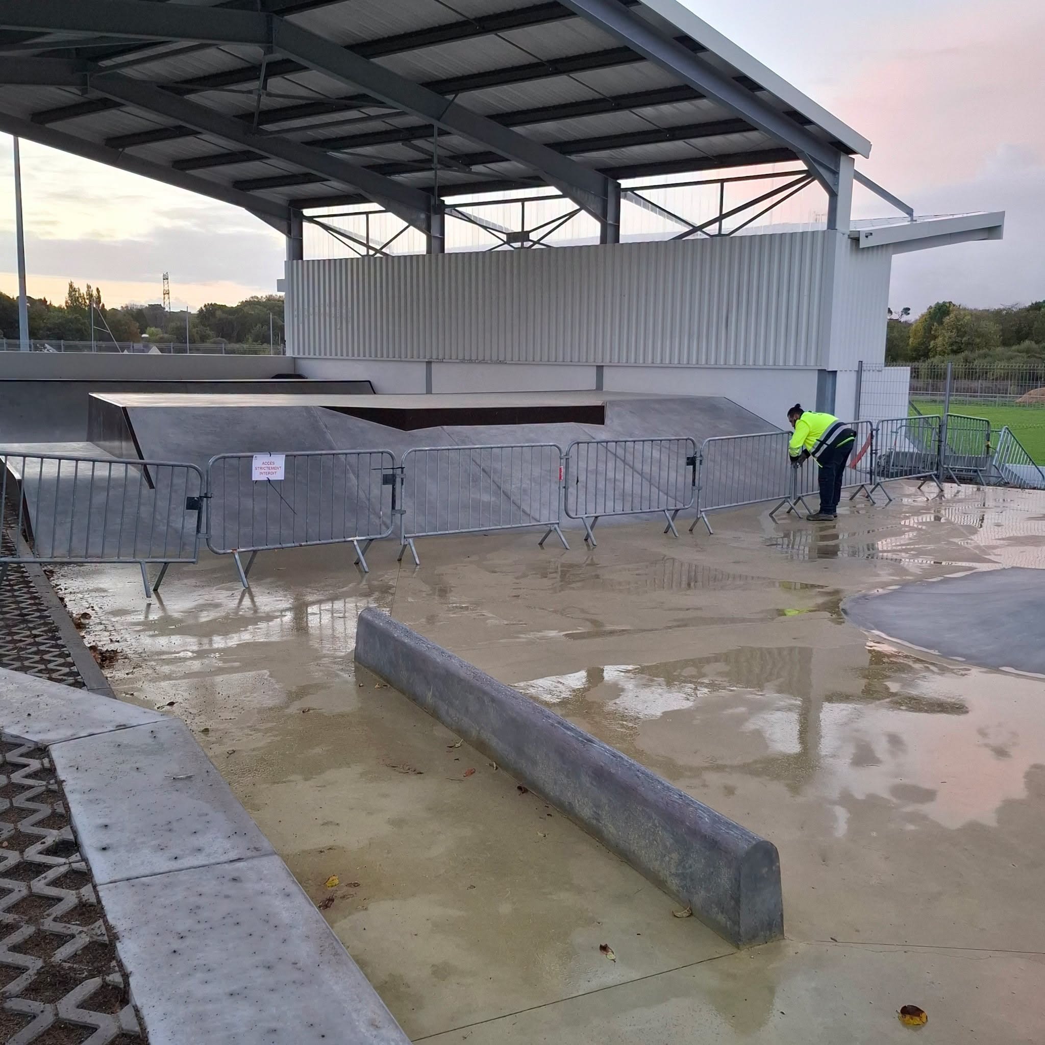 Photographie d’un skatepark couvert à Guidel, avec une rampe en béton et des barrières métalliques interdisant temporairement l’accès à certains modules. Le sol est mouillé et une personne en tenue de sécurité intervient sur place.